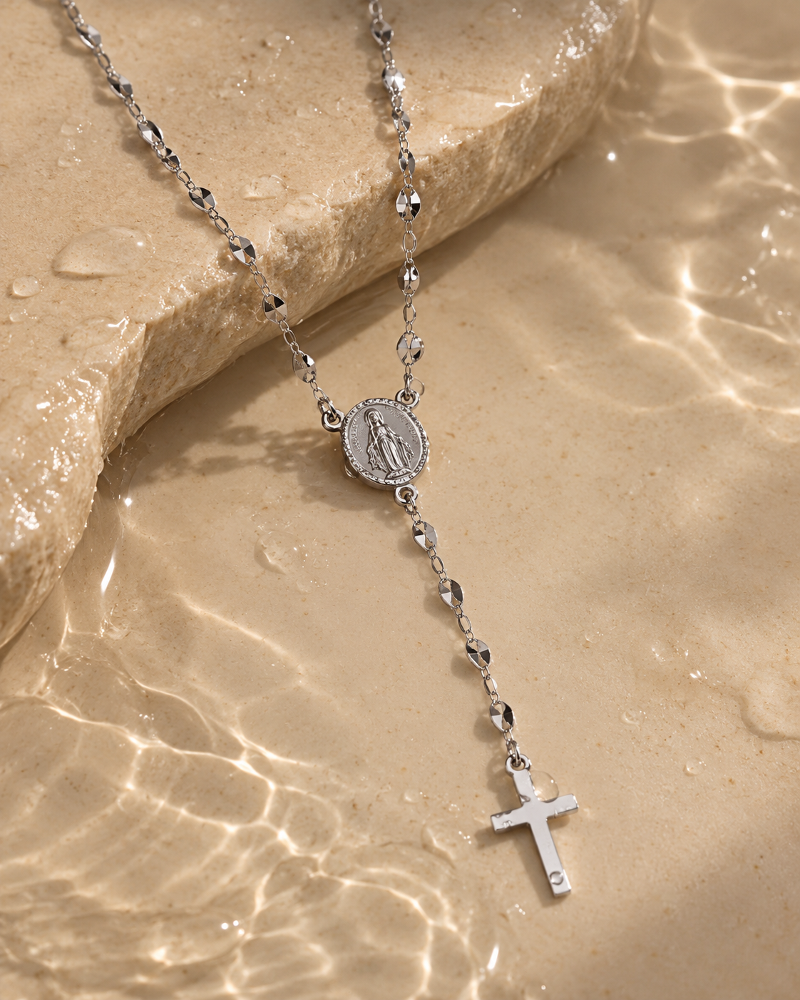 Collar tipo rosario de tono plateado con medalla ovalada y cruz, sobre piedra junto al agua con reflejos de luz.